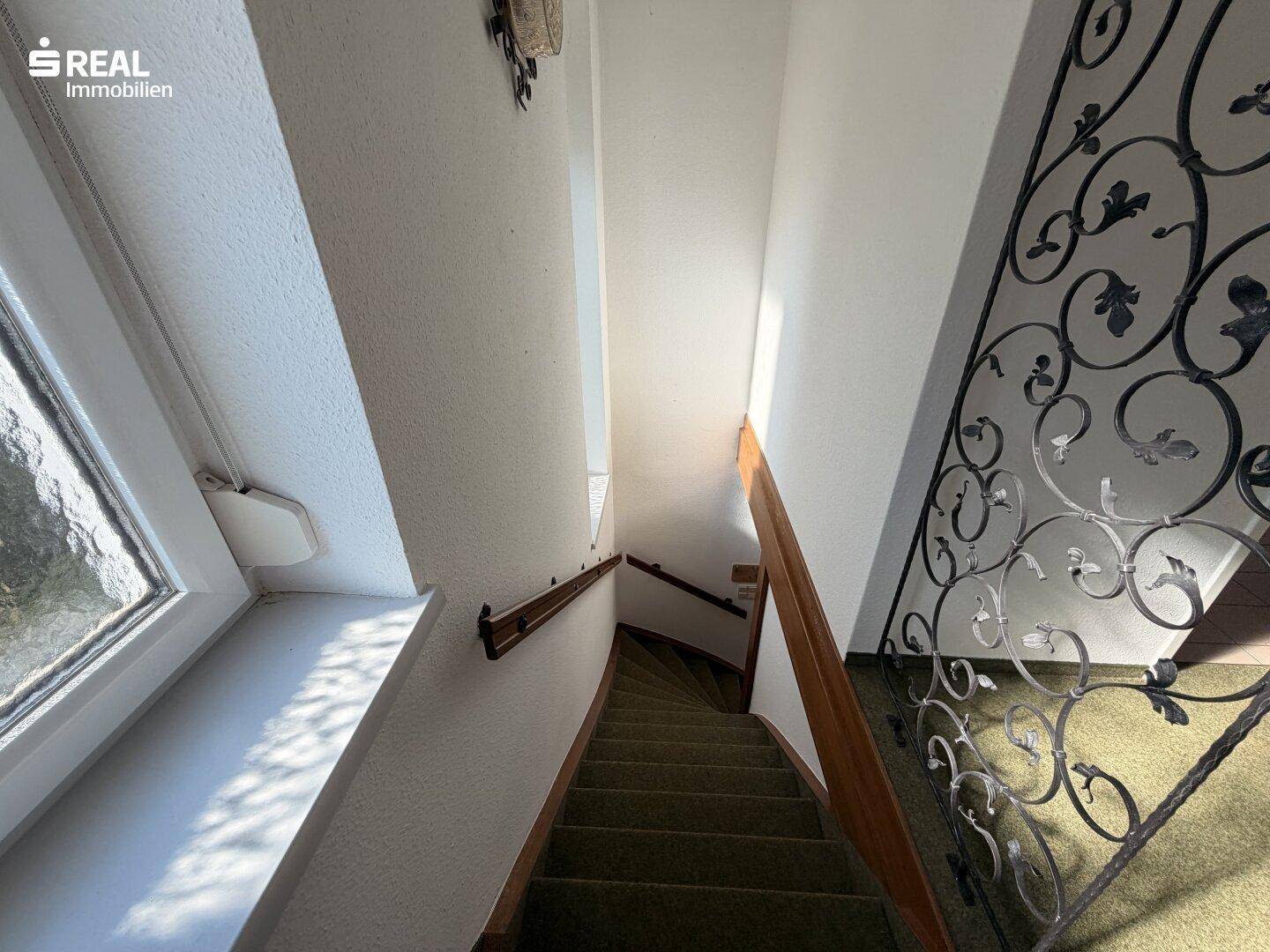 Helles Treppenhaus mit Teppichboden, Holzhandlauf und dekorativem Geländer.