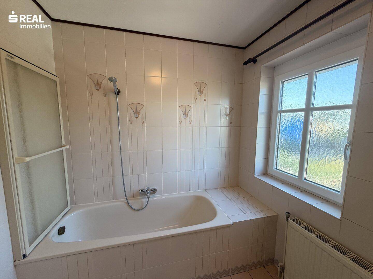 Helles Badezimmer mit Badewanne, Duschvorrichtung und Fenster, ausgestattet mit weißen Fliesen.