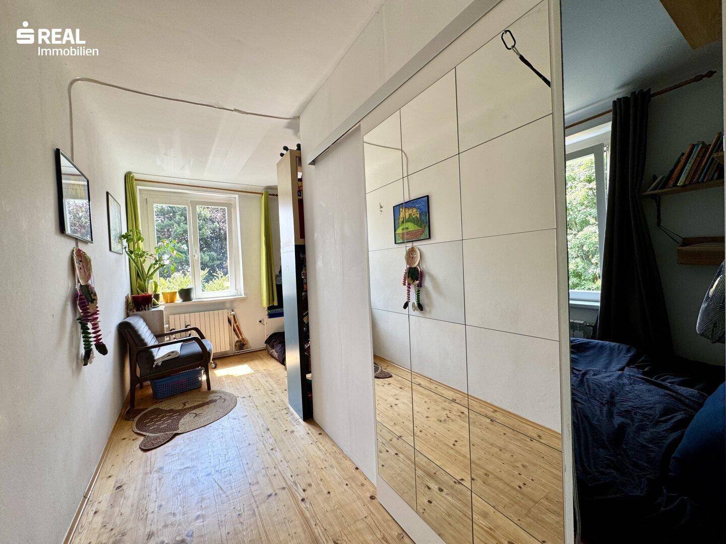 Schlafzimmer mit Holzdielenboden, Spiegelschiebetür und Fenster mit Blick ins Grüne.