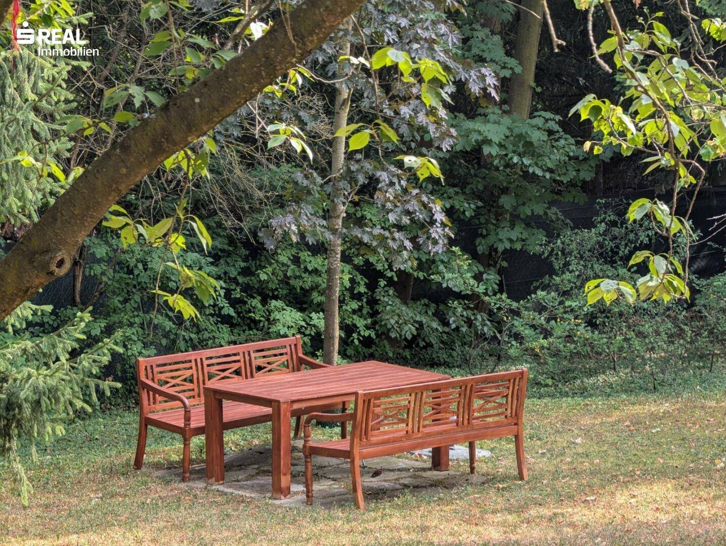 Gemütlicher Sitzbereich im Garten mit Holztisch und Bänken unter schattenspendenden Bäumen.