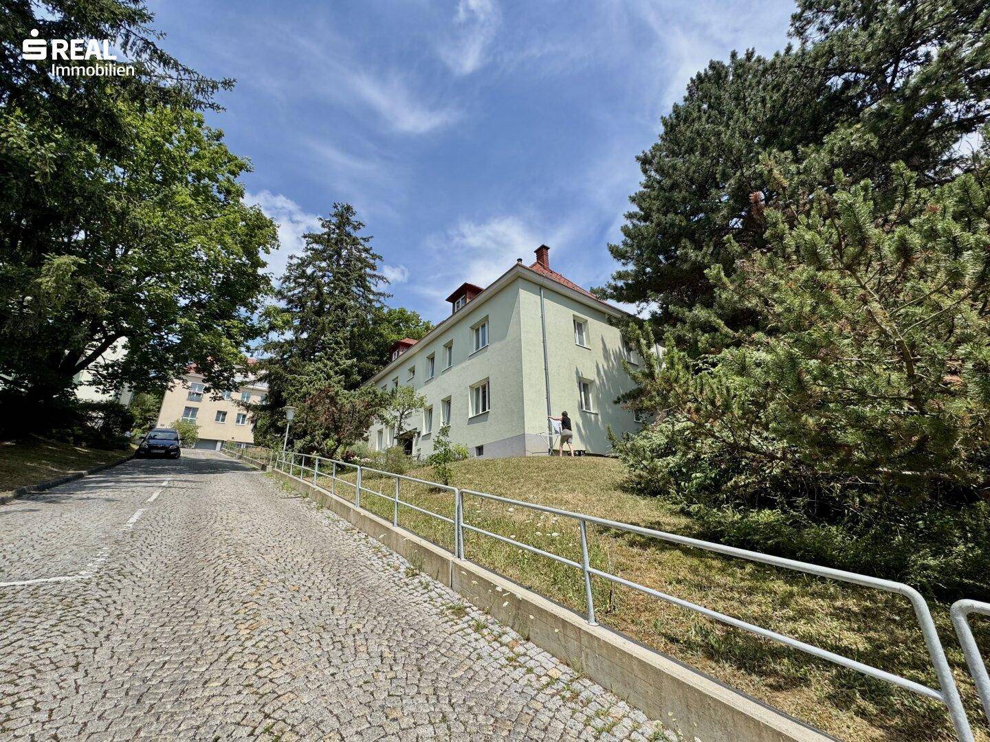 Mehrfamilienhaus mit heller Fassade, umgeben von Bäumen und einer gepflasterten Zufahrt.