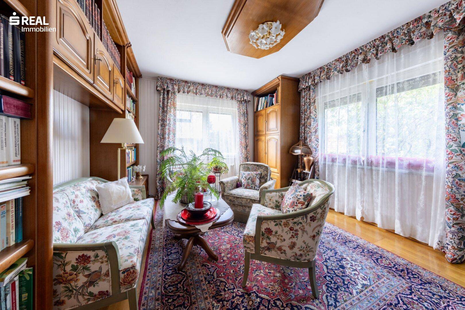 Gemütliches Wohnzimmer mit floralen Möbeln, Bücherregalen und traditionellem Teppich.
