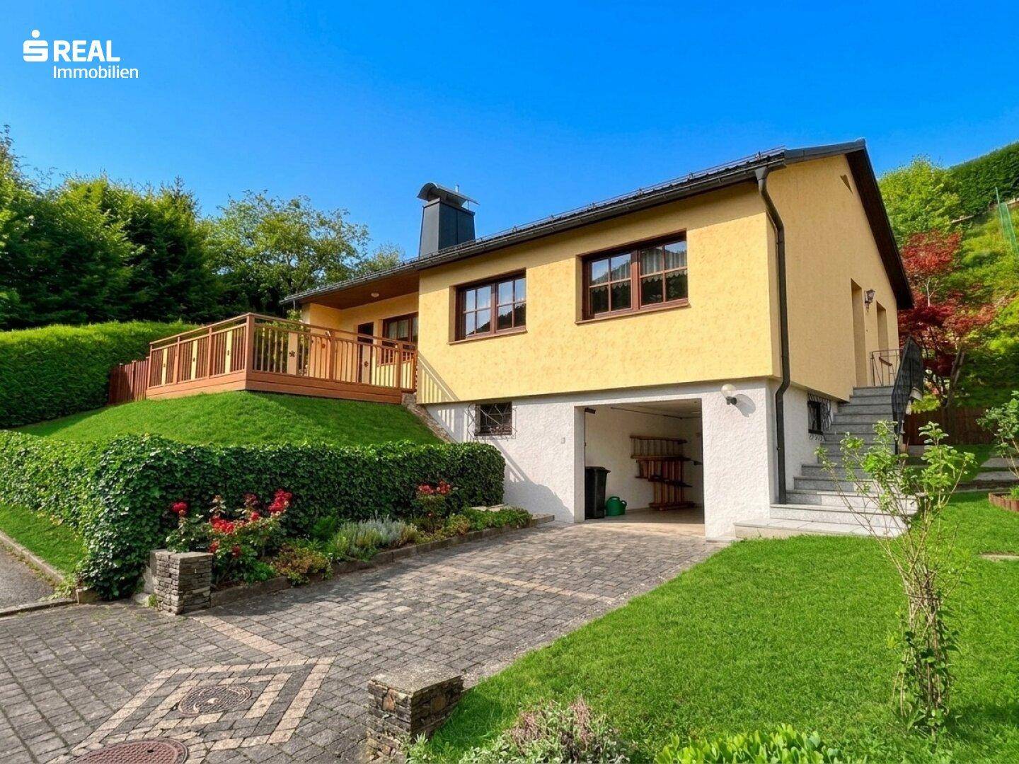 Vorderansicht des Hauses mit integrierter Garage, gepflasterter Zufahrt und erhöhter Holzterrasse.