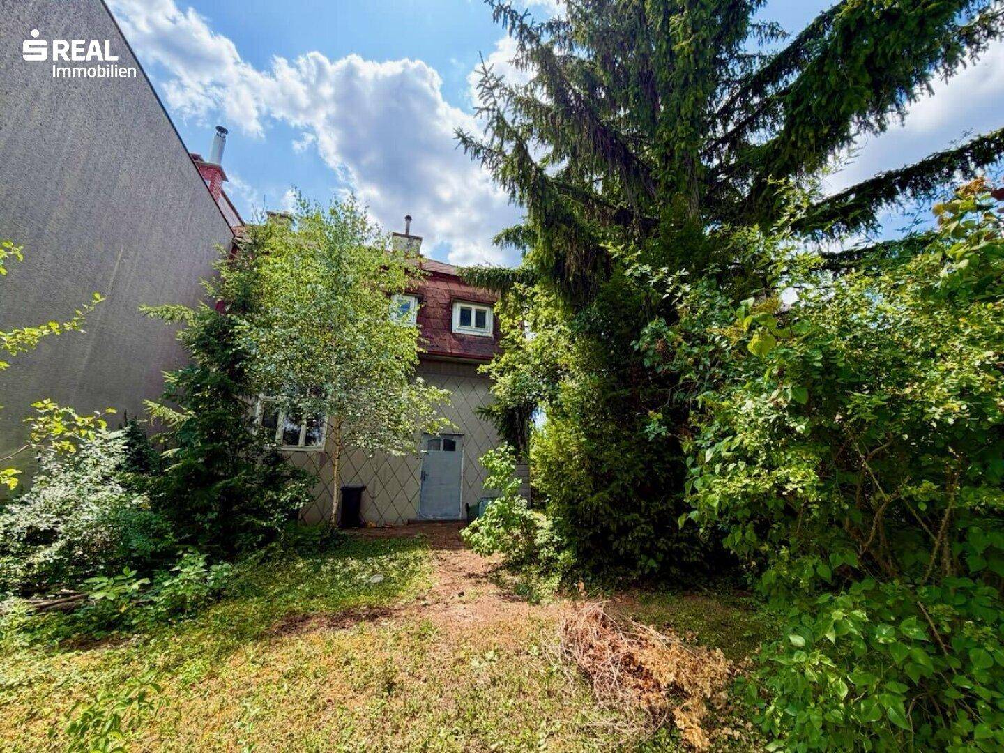 Rückseite des Hauses, umgeben von dichtem Grün und Bäumen, mit einem Zugang zum Garten.