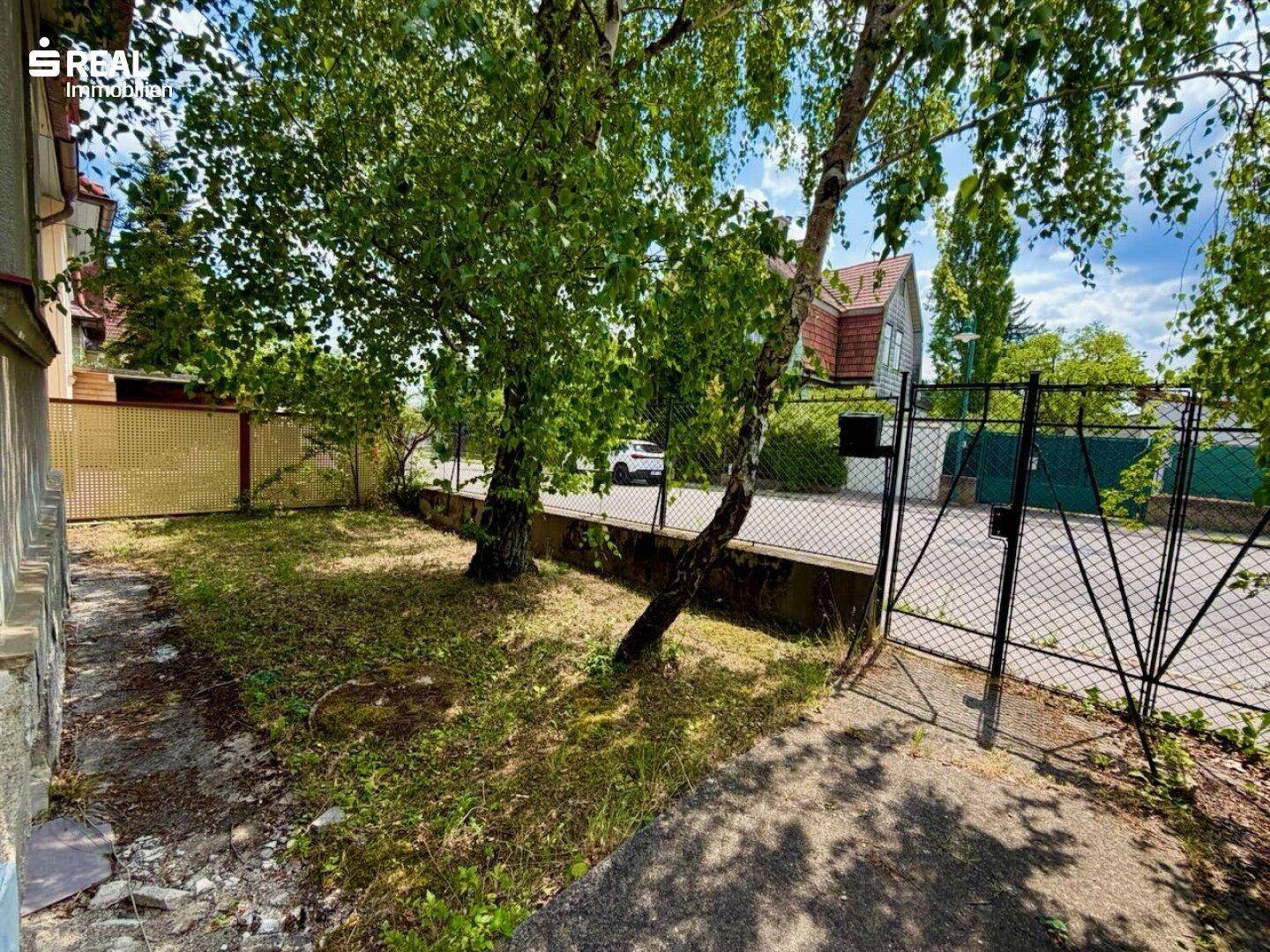 Seitlicher Blick auf das Grundstück mit Zaun, Tor und Zugang zur Straße, umgeben von Bäumen.