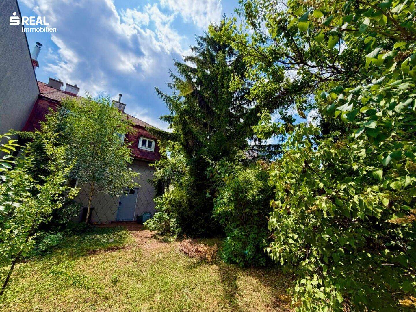 Ansicht der Hausrückseite vom Garten aus, umgeben von üppiger Vegetation und hohen Bäumen.