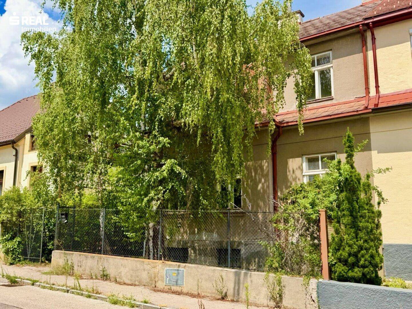 Ansicht der Hausfassade von der Straße, umgeben von Bäumen und einem Zaun, mit weiteren Gebäuden im Hintergrund.