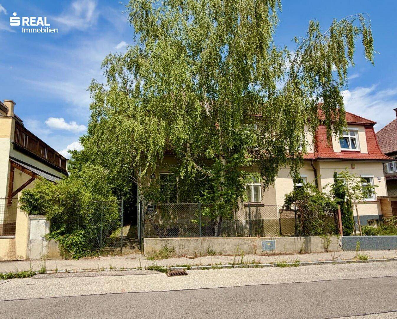 Vorderansicht des Hauses von der Straße aus, teilweise verdeckt von einem großen Baum und einem Zaun.
