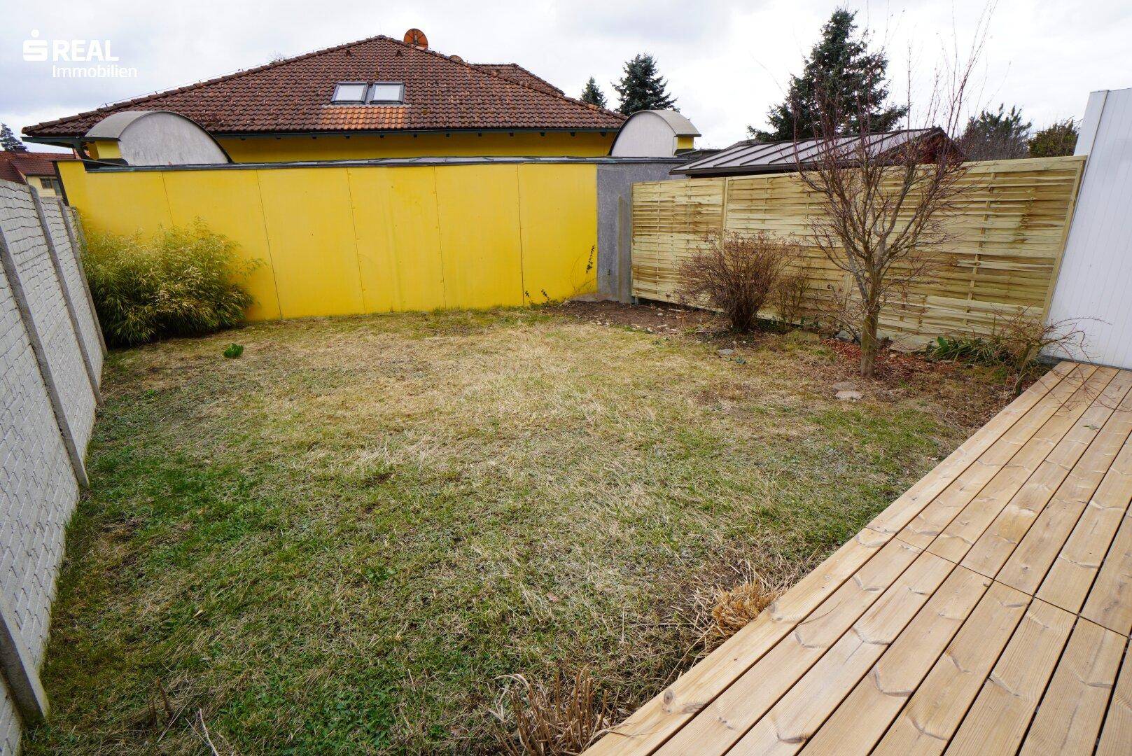 Kleiner Garten mit Rasenfläche und Holzterrasse, umgeben von Zäunen.