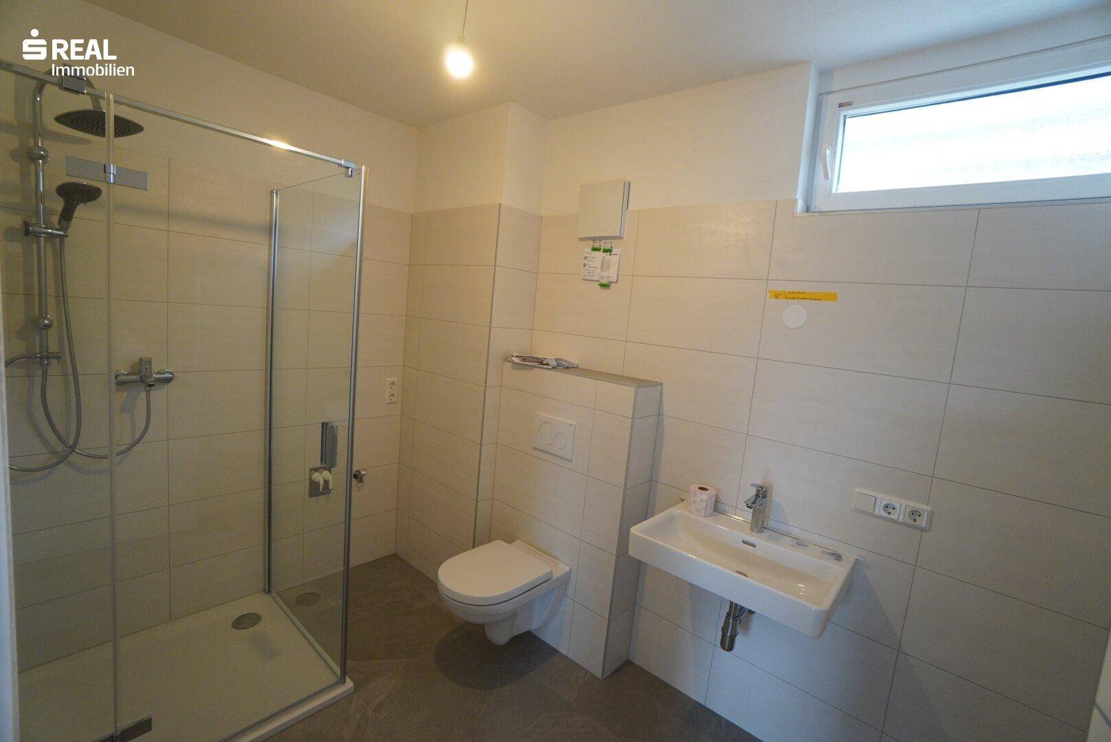 Helles Badezimmer mit Dusche, WC, Waschbecken und Fenster.