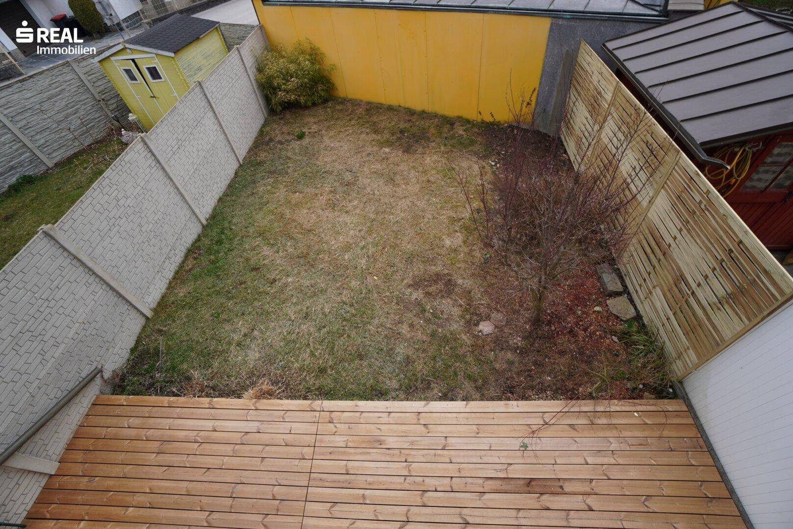 Kleiner Gartenbereich mit Holzterrasse und Rasenfläche, umgeben von Zäunen und einem Schuppen.