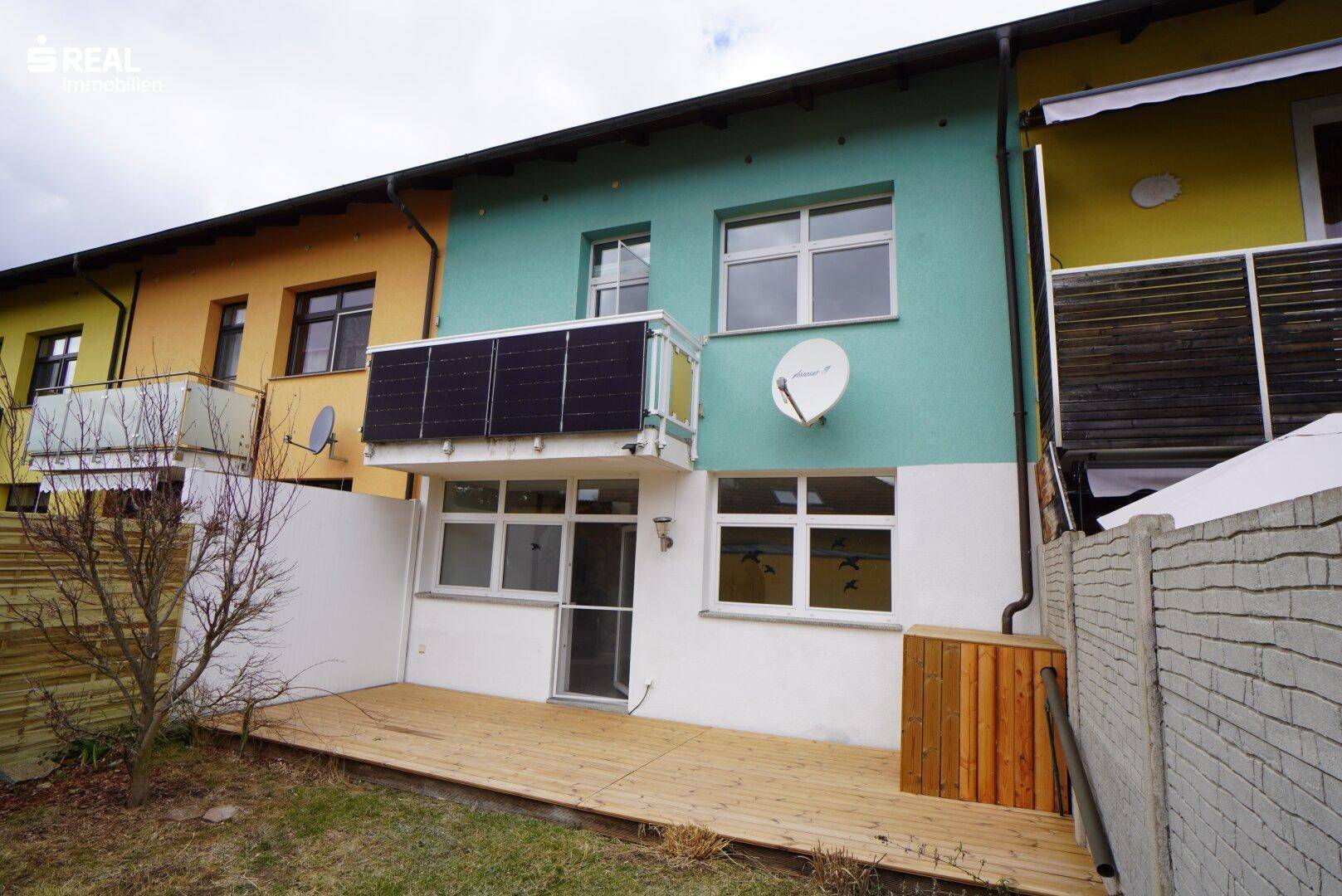Rückseite eines Reihenhauses mit Holzterrasse, kleinem Garten und Solarpaneelen am Balkon.