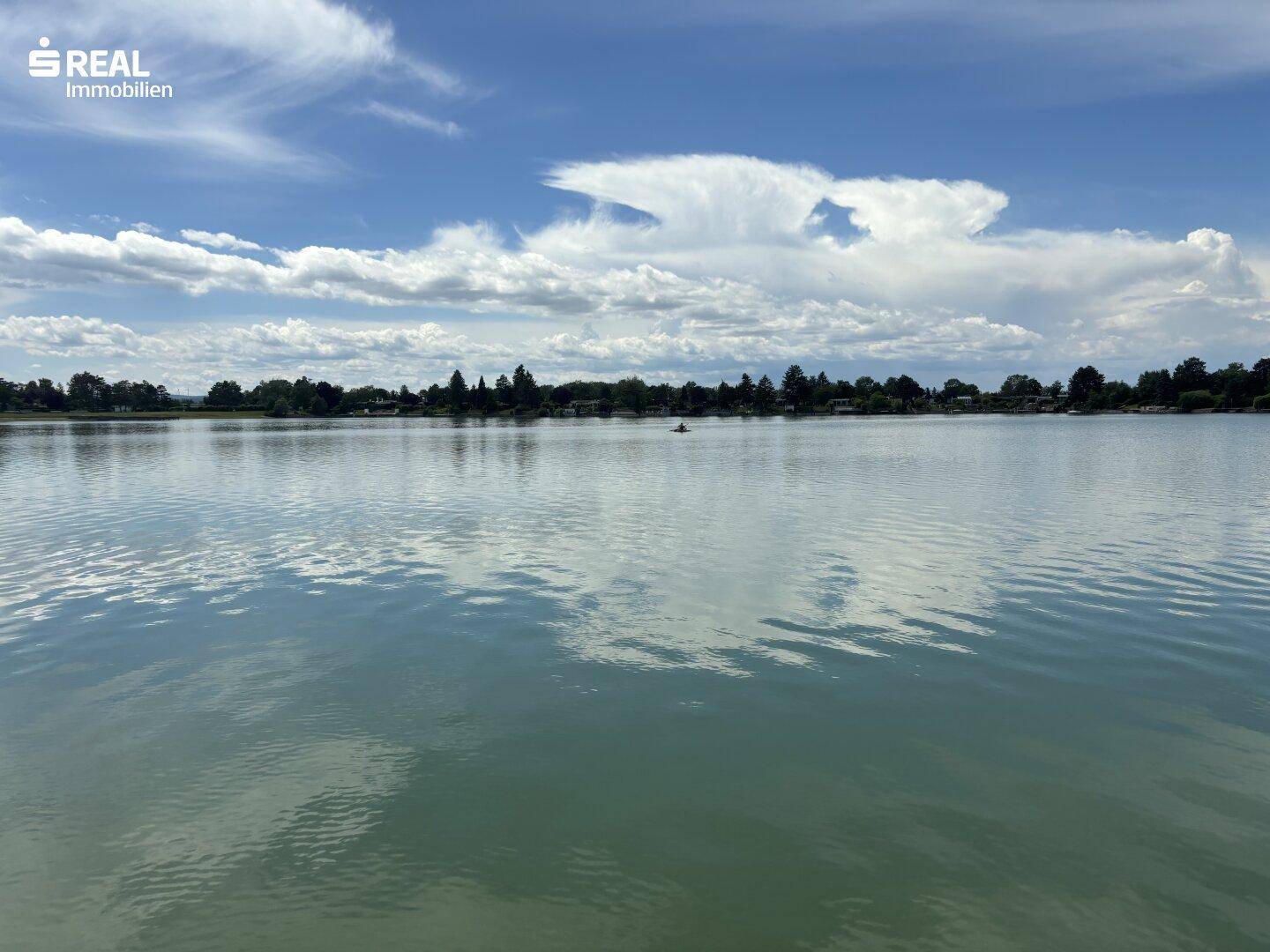 Weitläufiger Blick über einen ruhigen See unter einem bewölkten Himmel mit grünen Ufern in der Ferne.