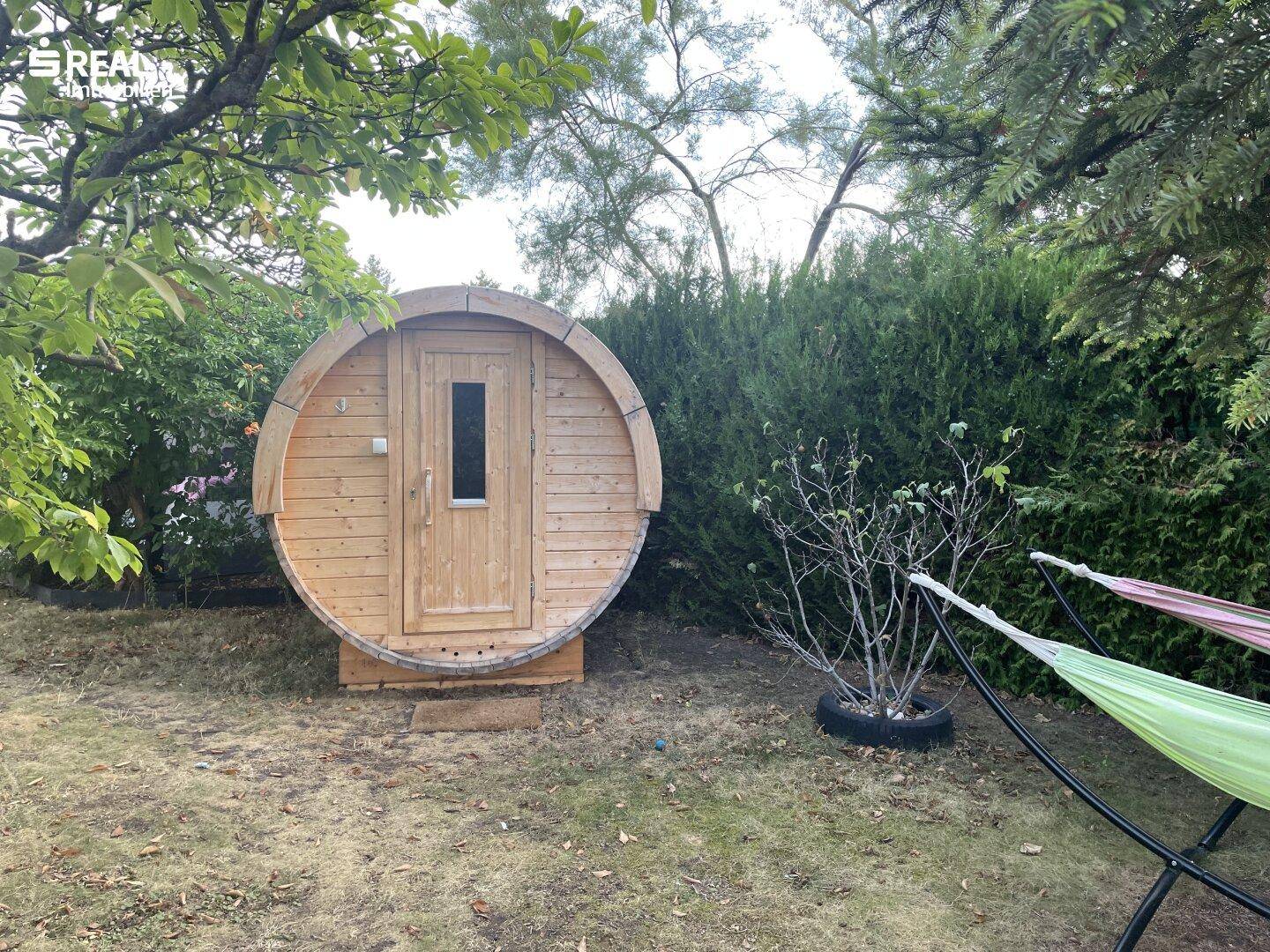 Fasssauna aus Holz im Garten, umgeben von einer Hecke und Hängematten für Entspannung.