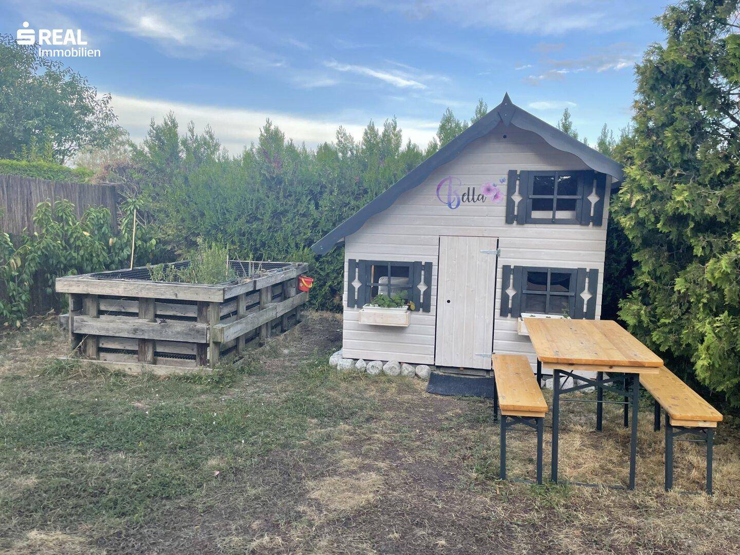 Kleines Holzspielhaus im Garten mit Hochbeet und rustikalem Picknicktisch, umgeben von Grün.