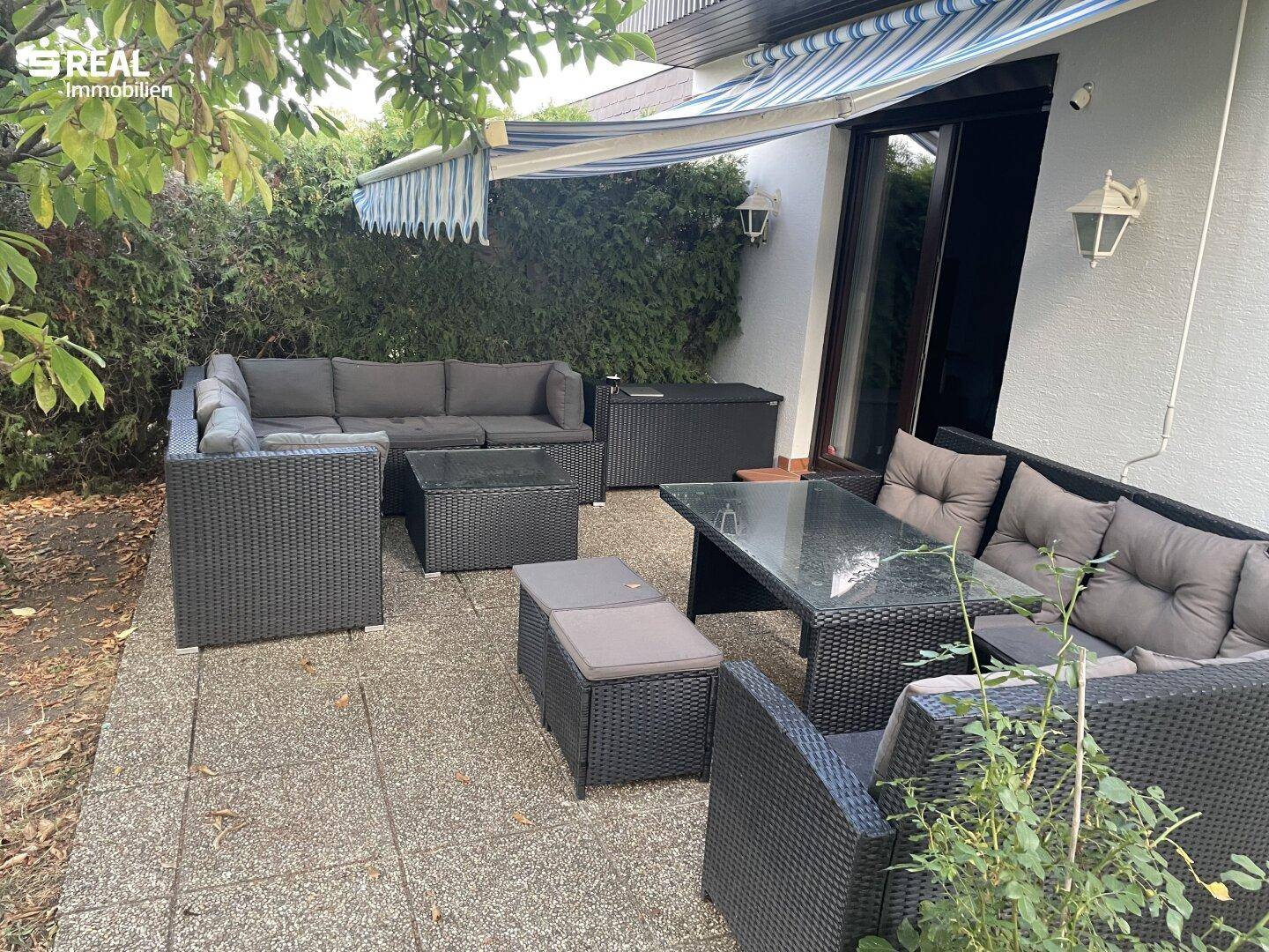 Möblierte Terrasse mit dunklen Rattanmöbeln, Glastisch und einer ausfahrbaren Markise für Schatten.