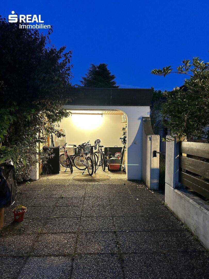 Einfahrt führt zu einer beleuchteten Garage mit Fahrrädern, umgeben von Bäumen in der Dämmerung.
