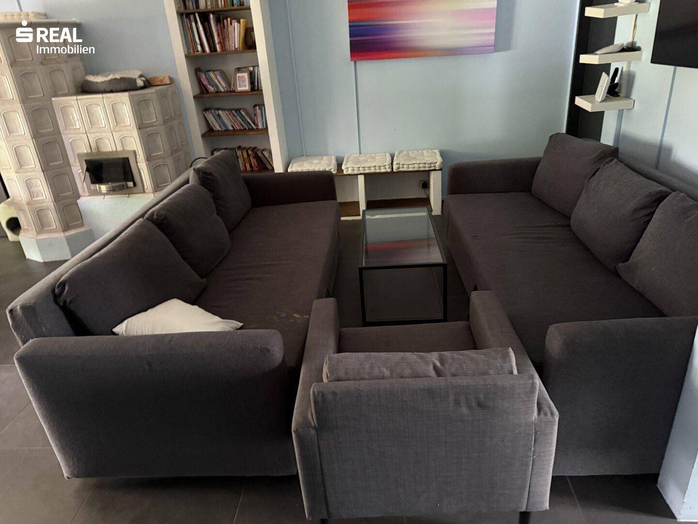 Wohnzimmer mit zwei dunklen Sofas, Sessel und Glastisch, ergänzt durch einen traditionellen Kachelofen.
