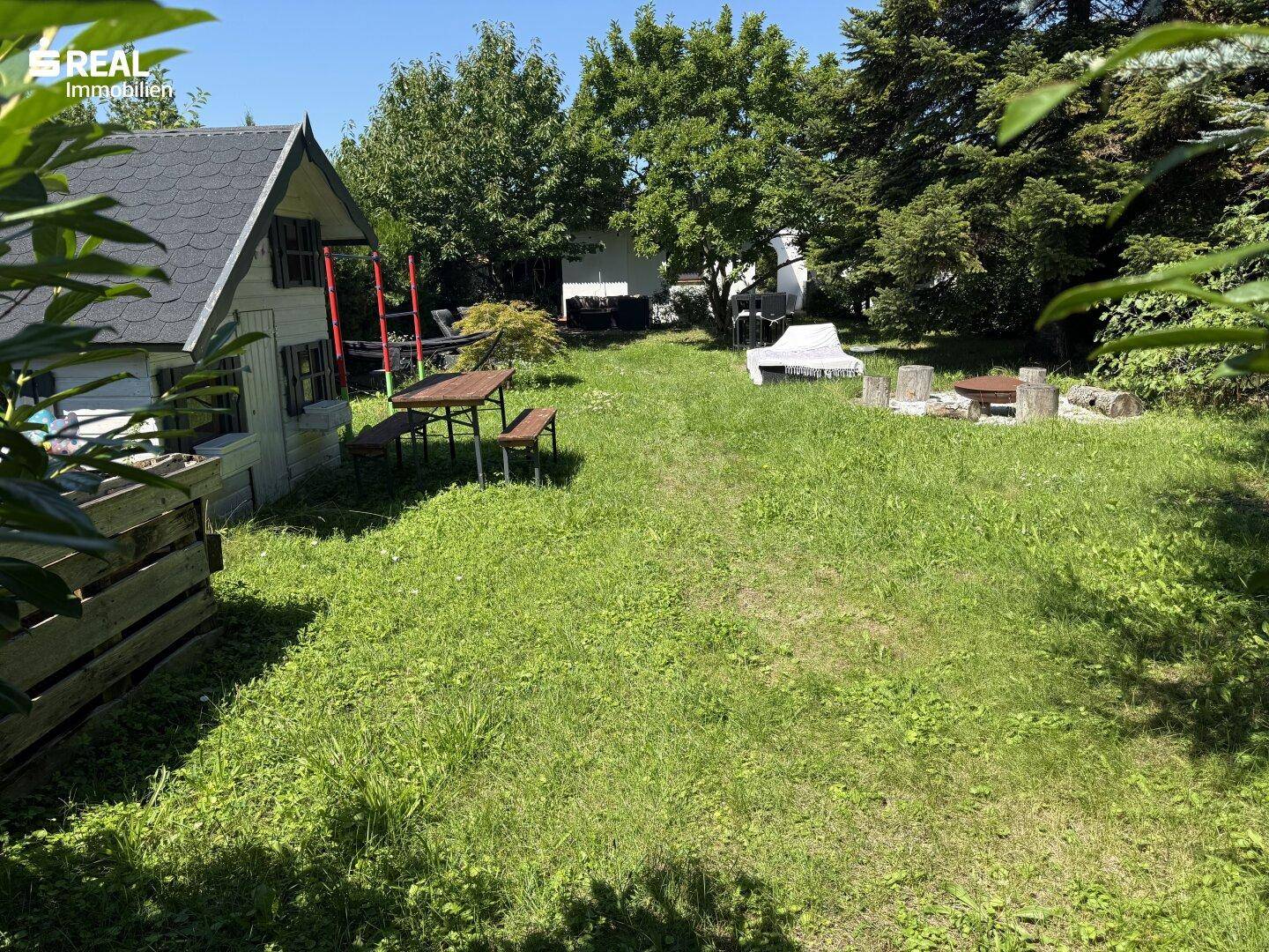 Gartenansicht mit Spielhaus, Picknicktisch und Feuerstelle auf einer sonnigen Rasenfläche.