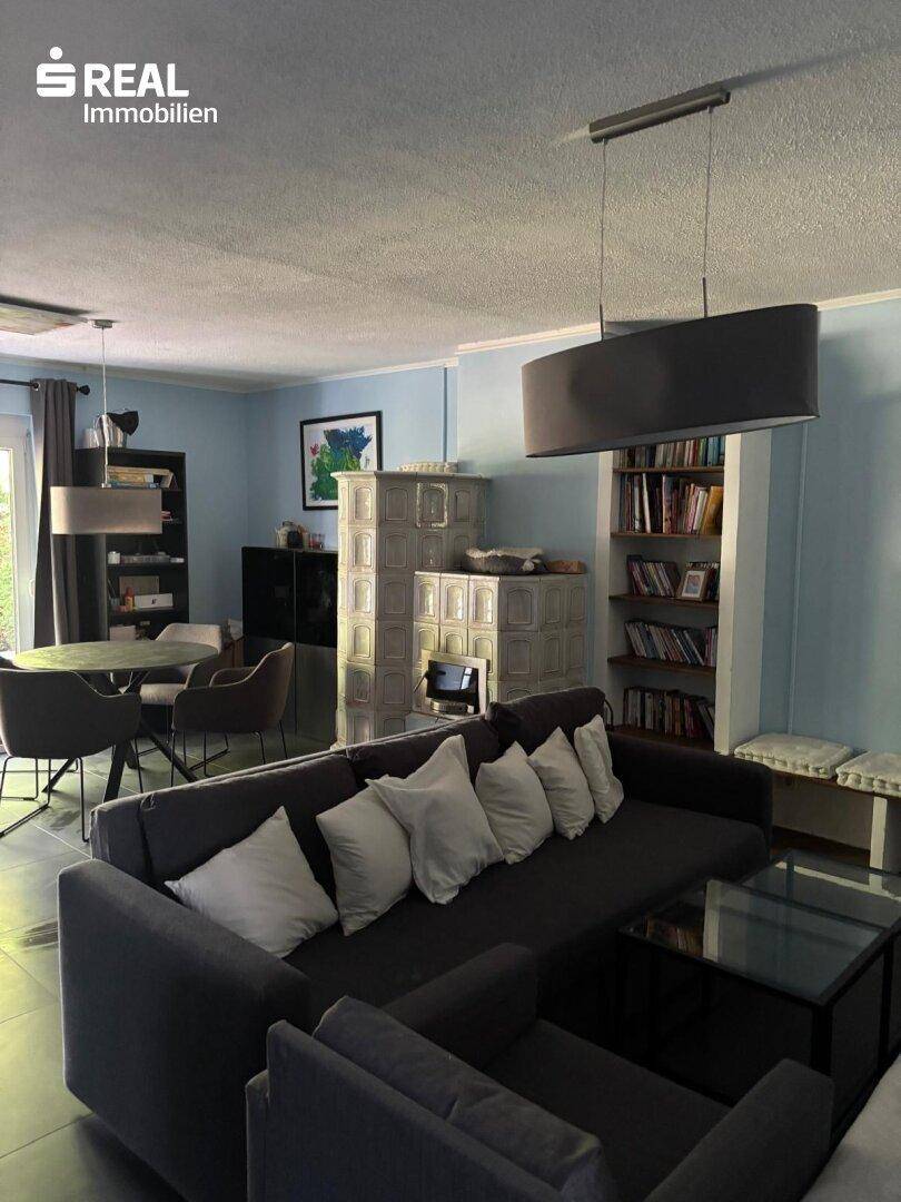 Geräumiges Wohnzimmer mit großem Sofa, Kachelofen und einem Essbereich im Hintergrund, hell und einladend.