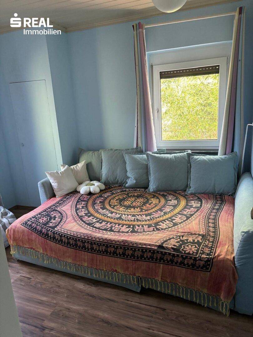 Gemütliches Schlafzimmer mit Tagesbett, farbenfroher Decke und einem Fenster mit Blick ins Grüne.