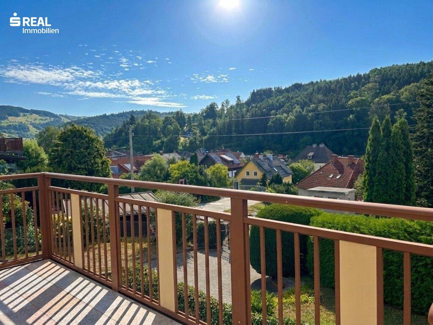 Sonniger Balkon mit weitem Blick über die umliegenden Häuser und die bewaldeten Berge.