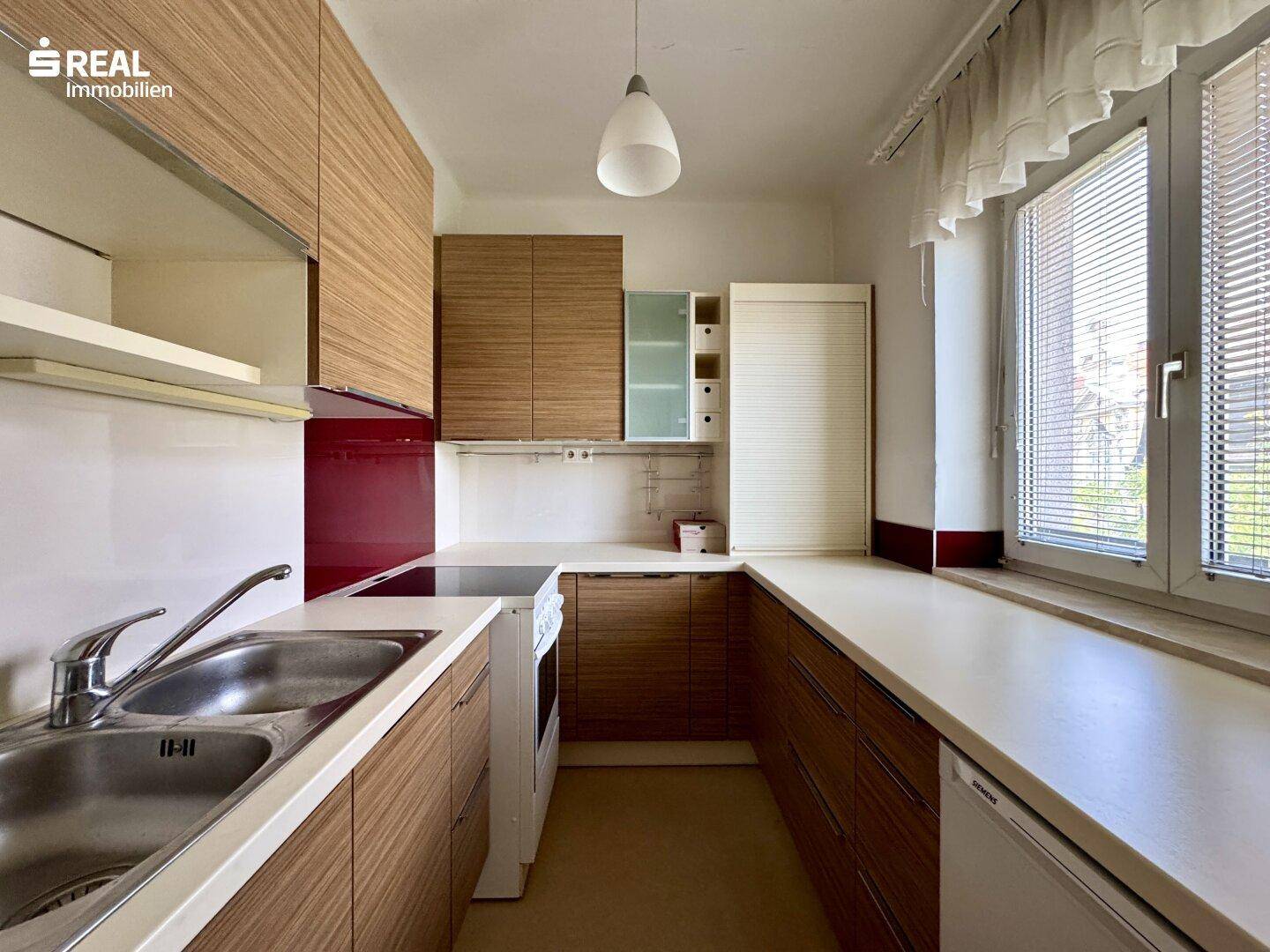 Funktionale Küche mit Holzfronten, Doppelspüle und Fenster mit Jalousien für natürliches Licht.