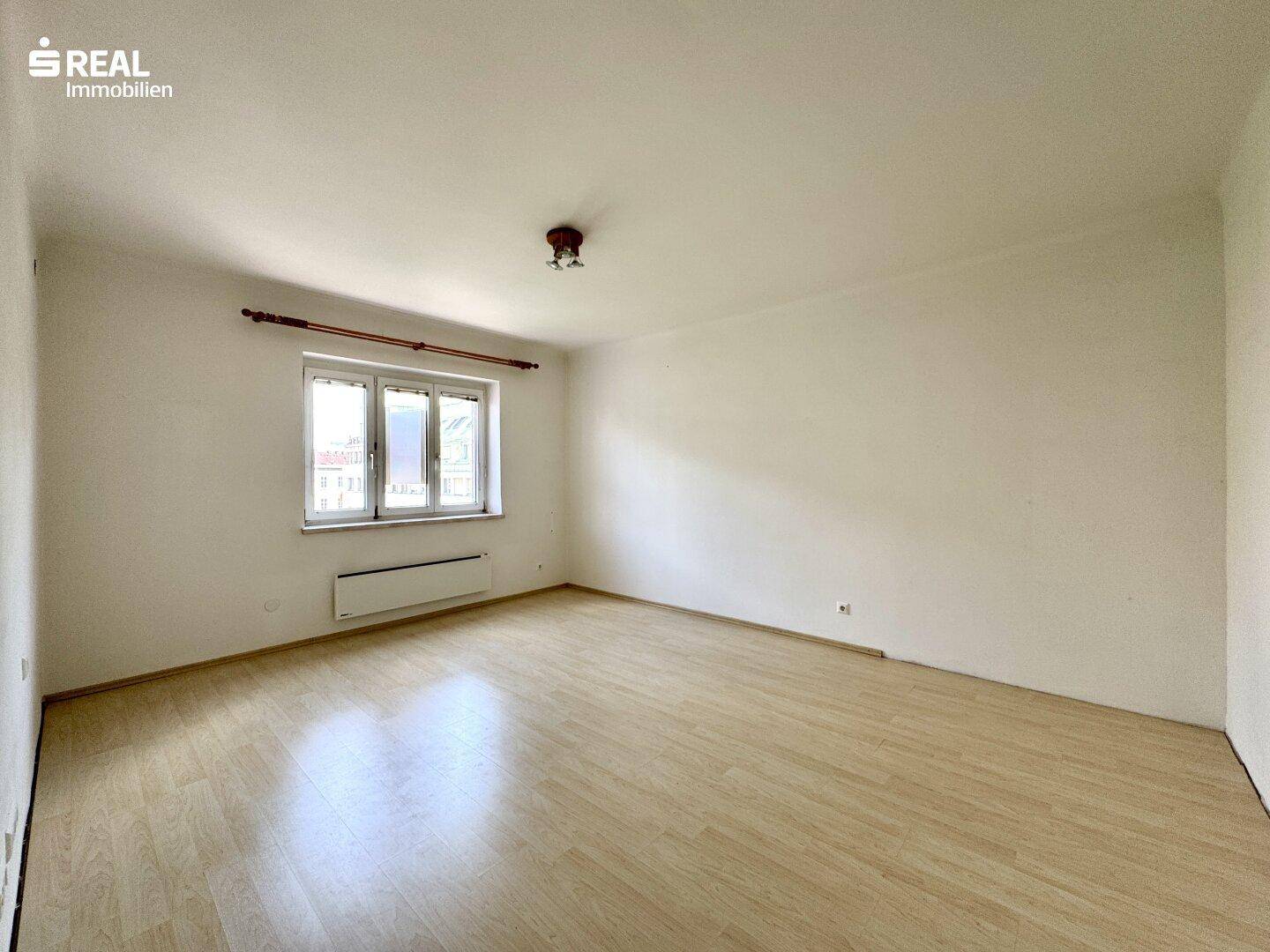 Helles, leeres Zimmer mit Laminatboden und großem Fenster, ideal für individuelle Gestaltung.