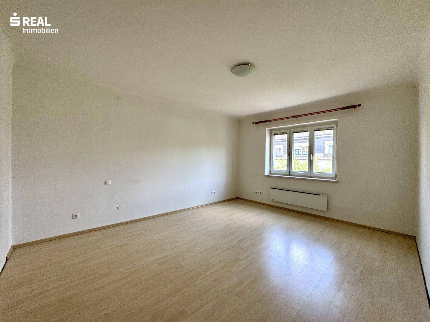 Geräumiges und helles Zimmer mit Laminatboden und Heizkörper unter dem Fenster.