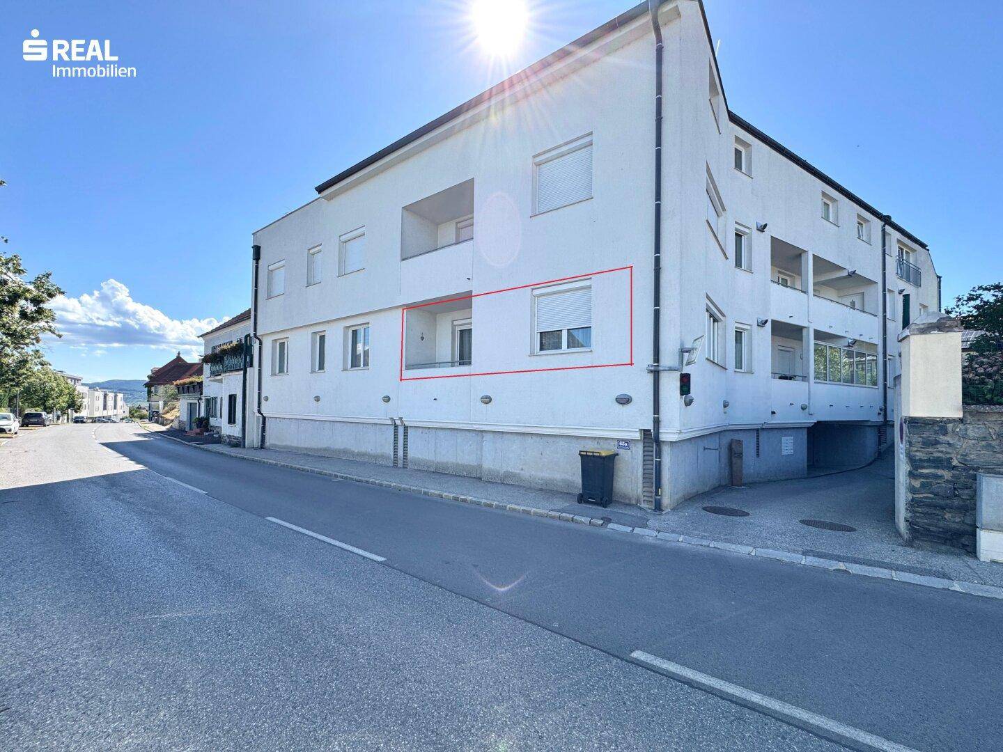 Modernes Mehrfamilienhaus an einer sonnigen Straße mit weißen Fassaden, Balkonen und Bergblick.