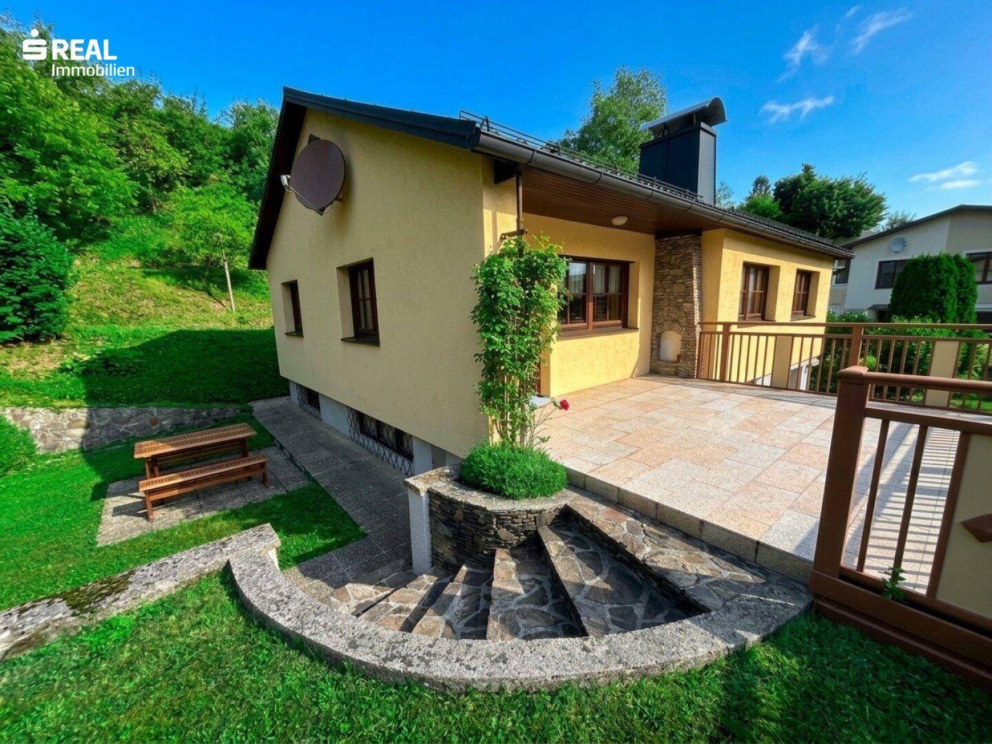 Seitenansicht des Hauses mit großer Steinterrasse und geschwungenen Stufen zum Gartenbereich.