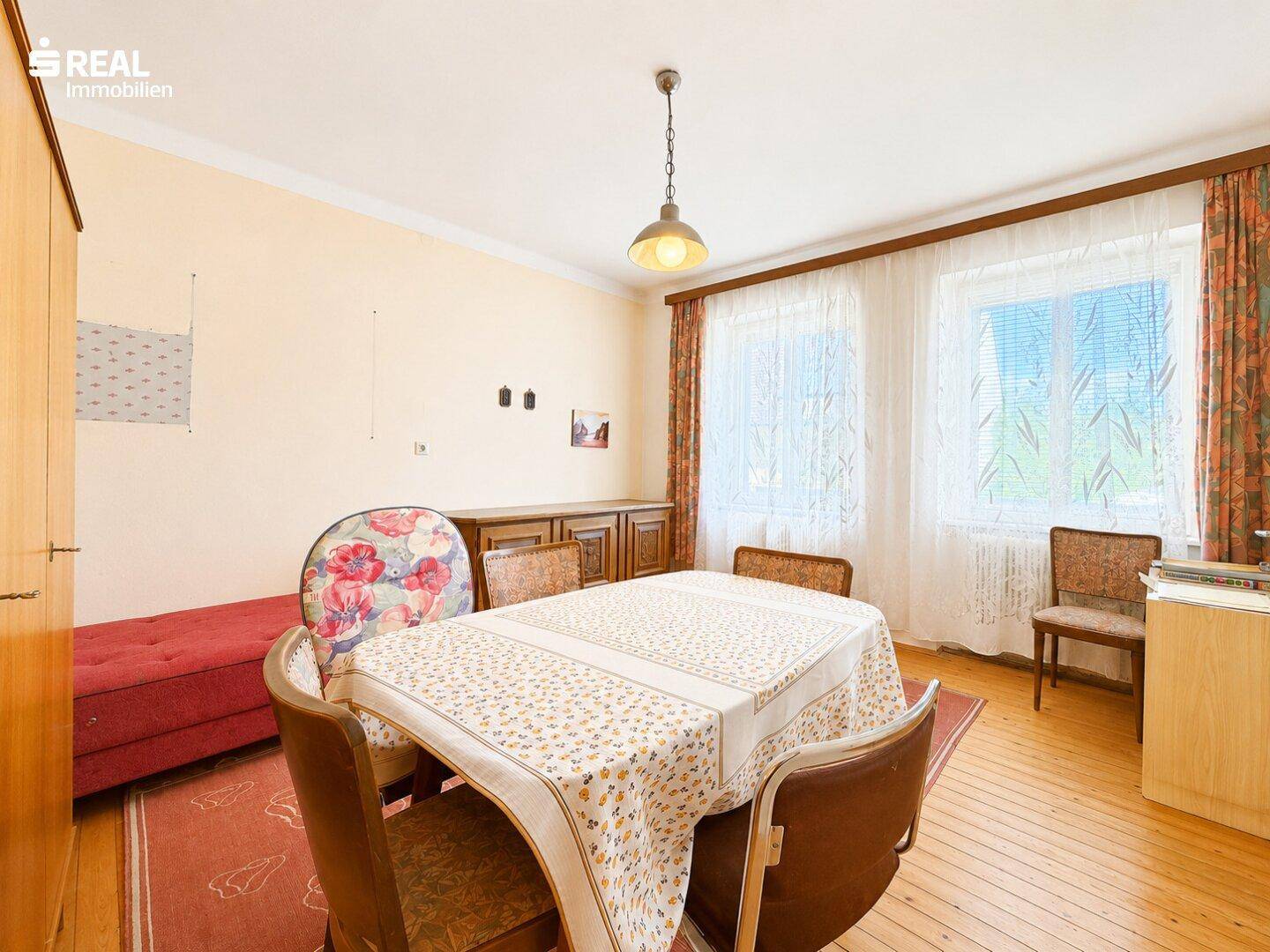 Helles Esszimmer mit großem Esstisch, Holzfußboden und Fenstern mit Jalousien.