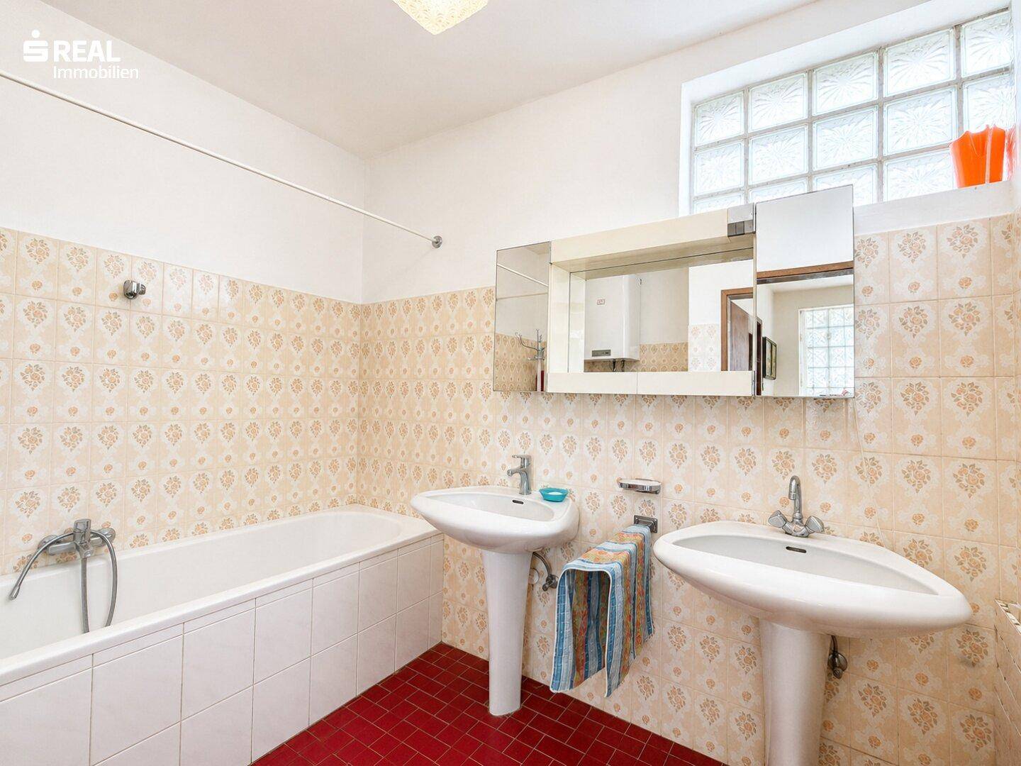 Badezimmer mit zwei Waschbecken, Badewanne und Glasbausteinfenster für Tageslicht.