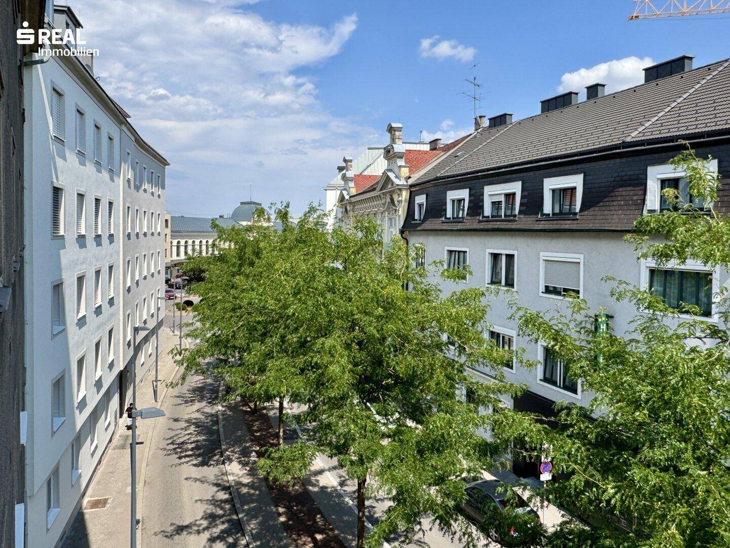 Blick auf eine belebte Stadtstraße mit historischen Gebäuden, grünen Bäumen und blauem Himmel.