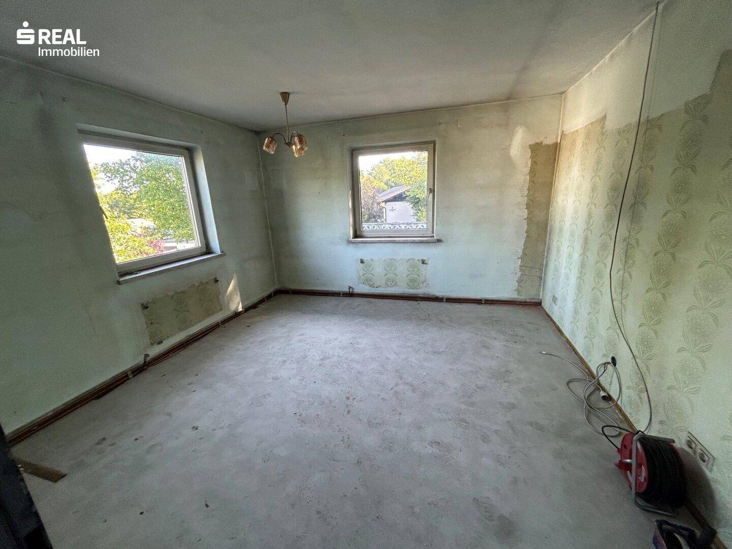 Geräumiges Zimmer in Renovierung mit zwei Fenstern und abgelöster Tapete.