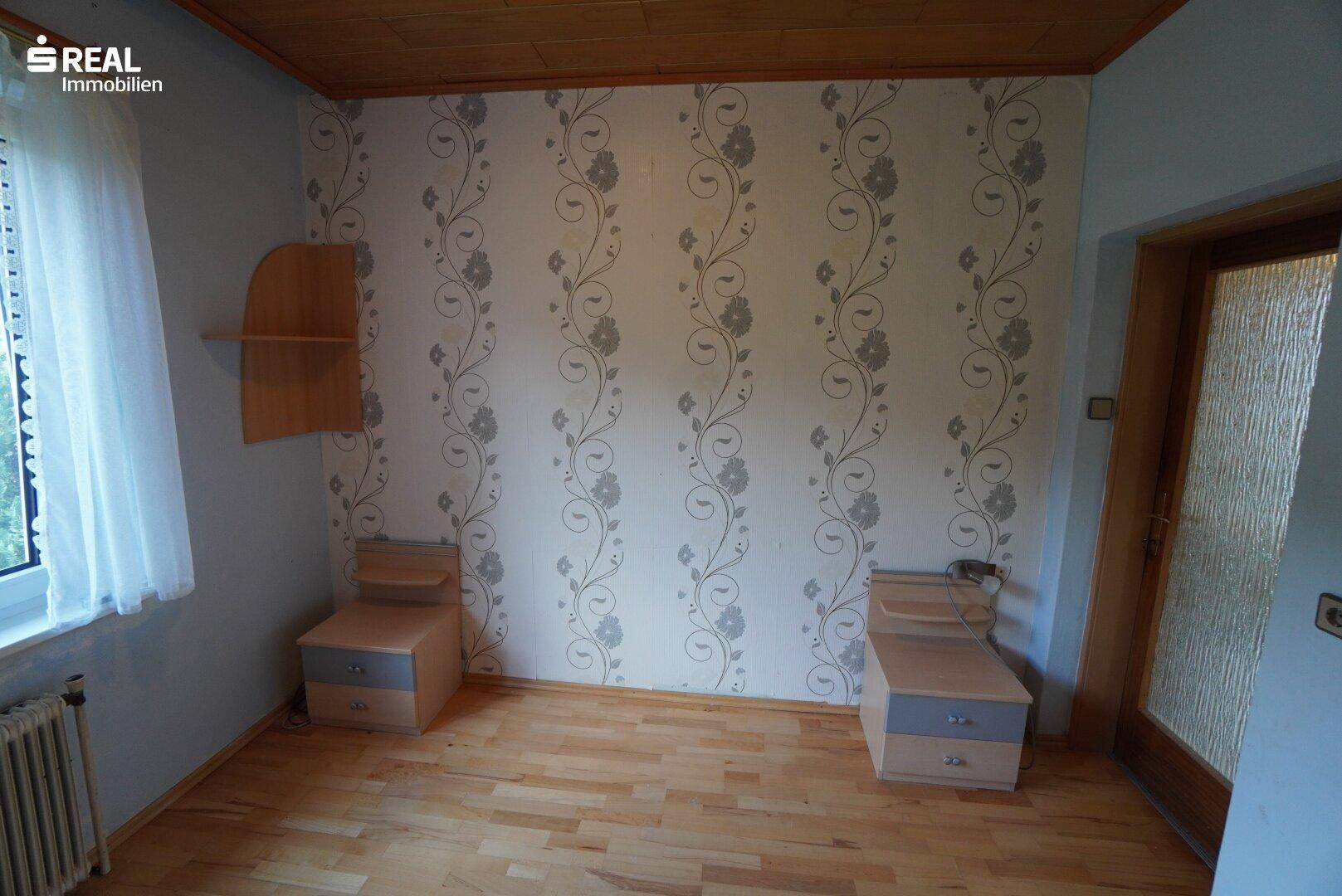 Schlafzimmer mit Parkettboden, Holzdecke und floraler Tapete an einer Wand.