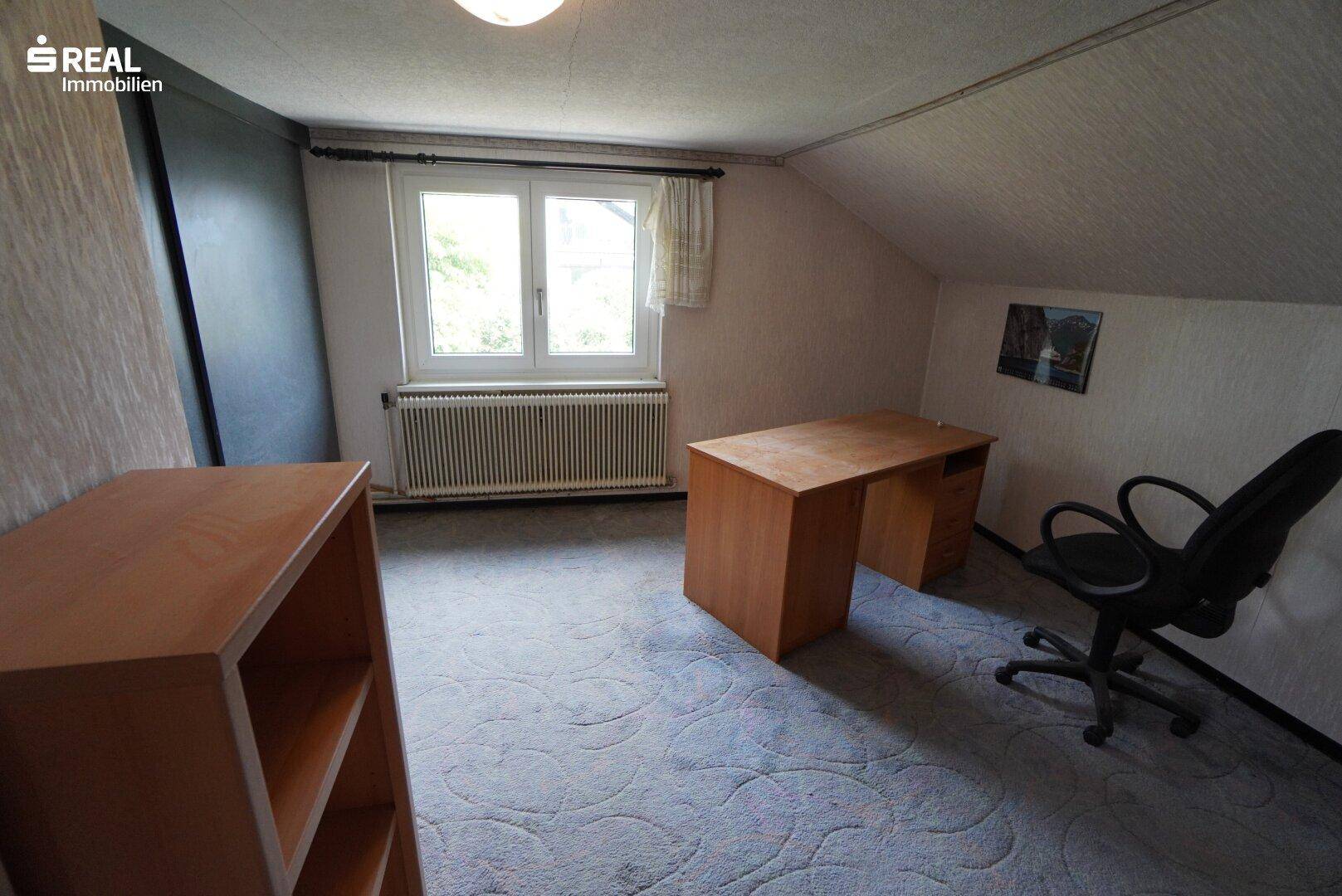 Dachgeschossbüro mit Teppichboden, Schreibtisch und Fenster mit Blick ins Grüne.