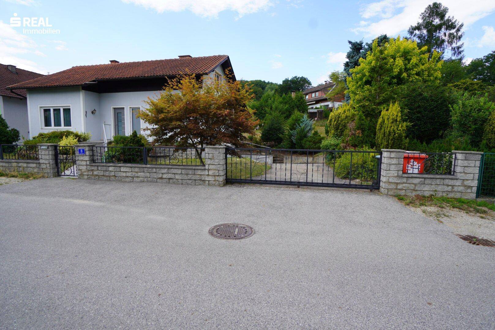Eingangsbereich des Grundstücks mit Zaun, Tor und Blick auf das Haus in grüner Umgebung.