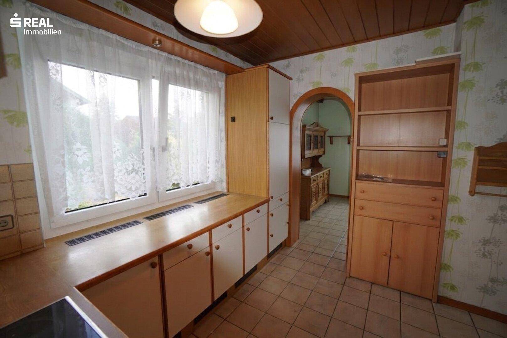 Küchenbereich mit großem Fenster, Holzdecke und integrierten Schränken, angrenzend an einen weiteren Raum.