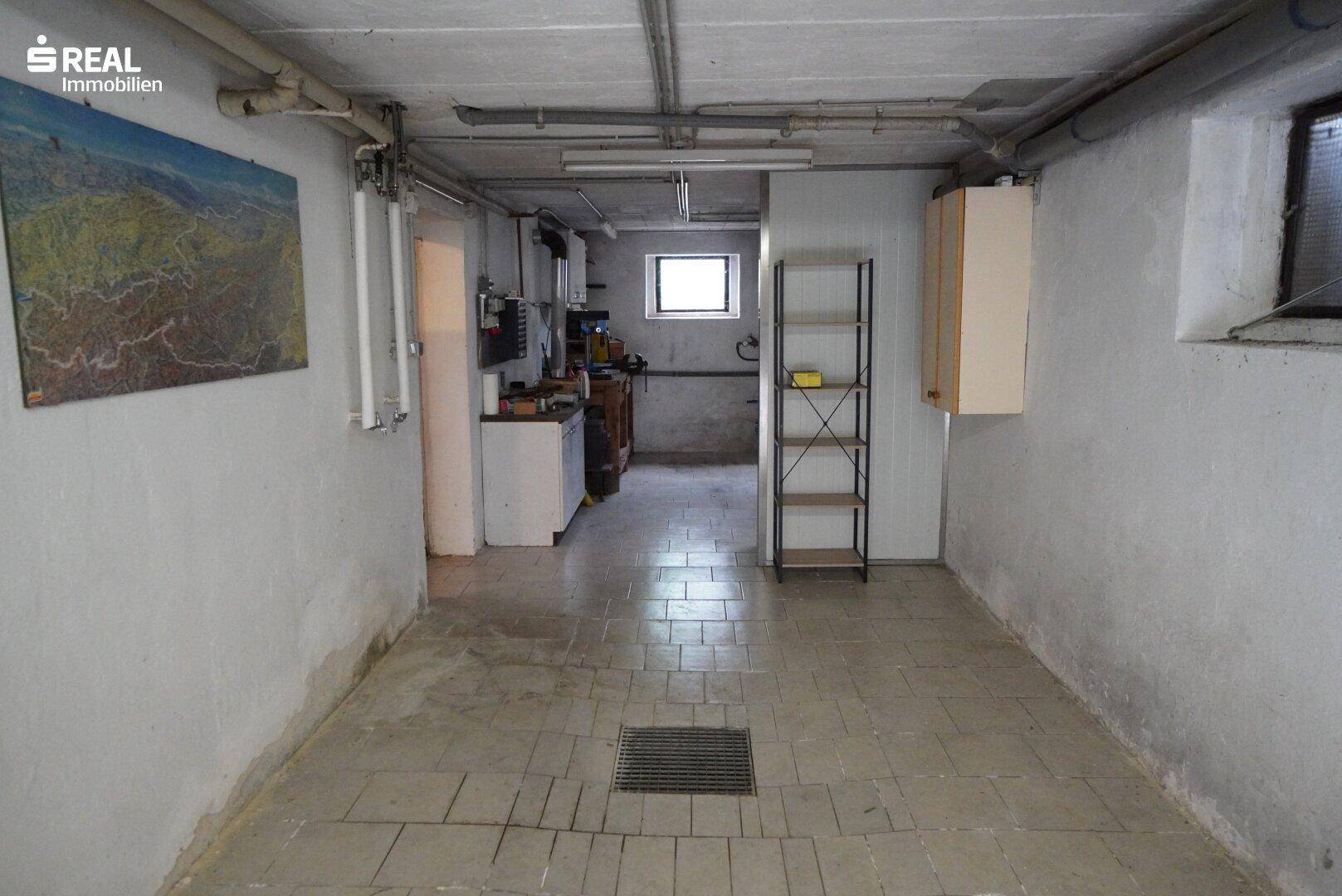 Langer Kellerraum mit Betonboden, kleinen Fenstern und einem Bereich für Werkstatt oder Lagerung.