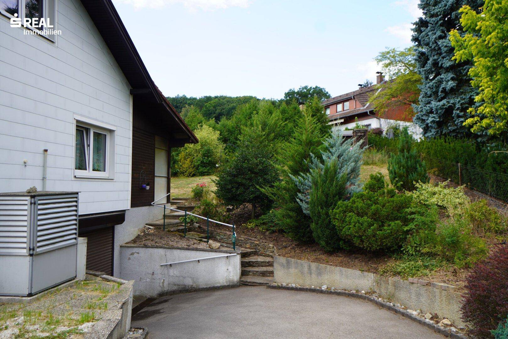 Seitenansicht des Hauses mit Zufahrt und üppiger Bepflanzung im Gartenbereich.