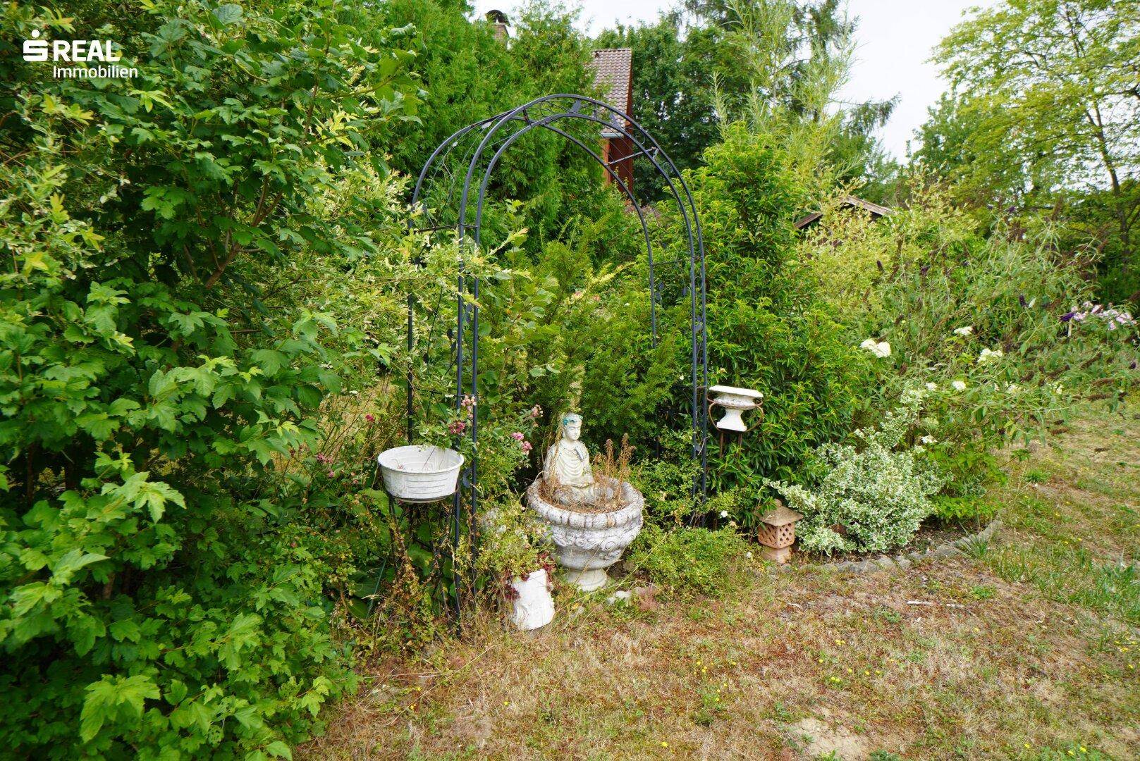 Dekorativer Gartenbereich mit einem Metallbogen, Statuen und üppiger Vegetation.