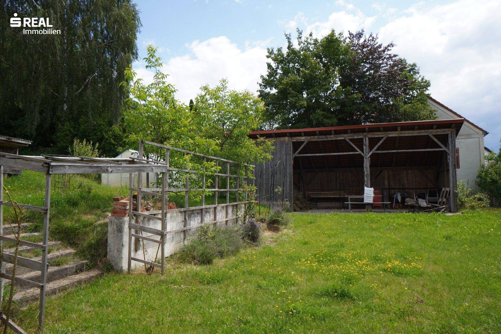 Grüner Gartenbereich mit Rasen, Bäumen und einem Holzunterstand im Hintergrund.