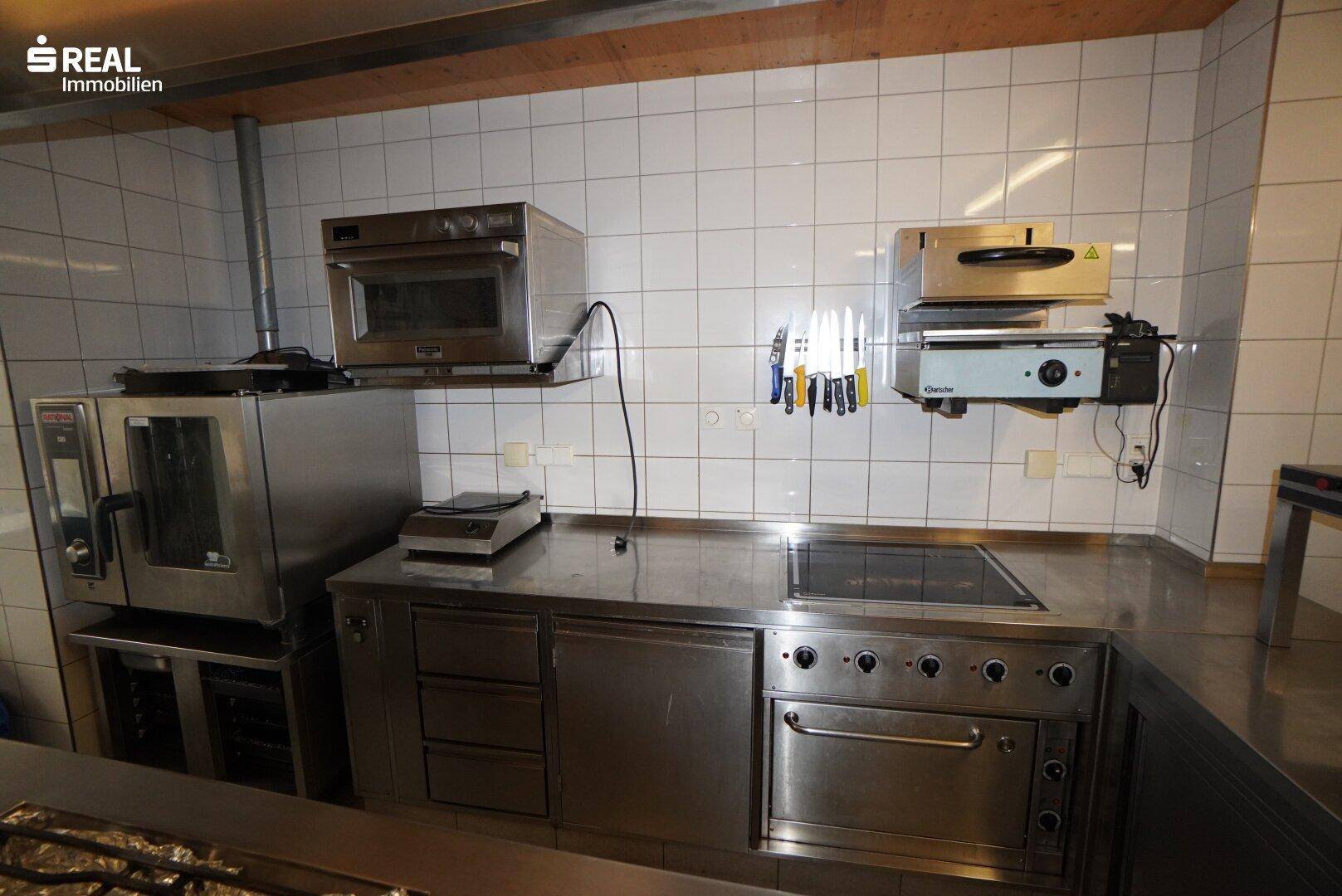 Detailansicht einer professionellen Küche mit Edelstahlgeräten, Backofen und weiß gefliesten Wänden.