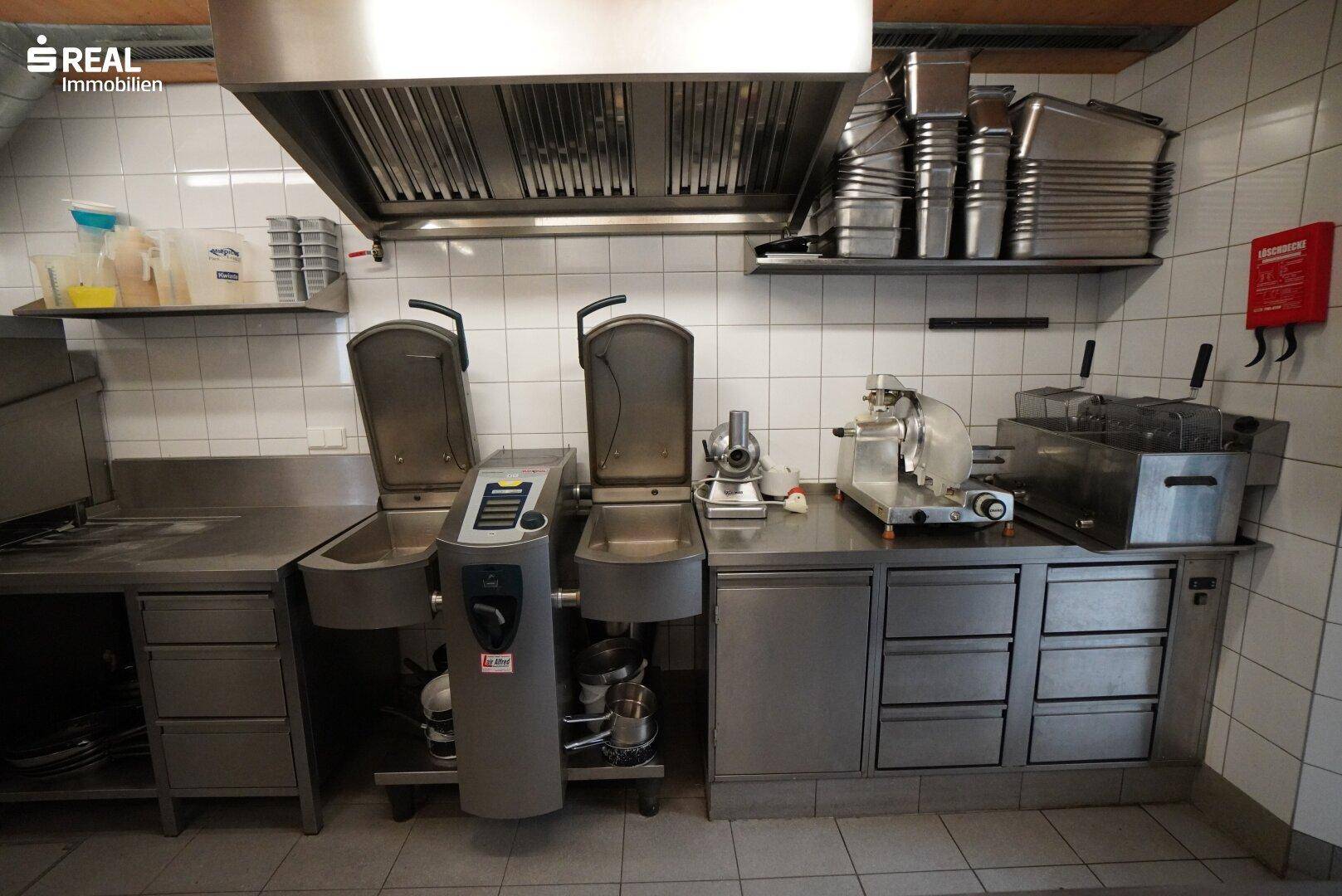 Weiterer Bereich der Großküche mit Edelstahlarbeitsflächen, Spülbecken und professionellen Geräten.