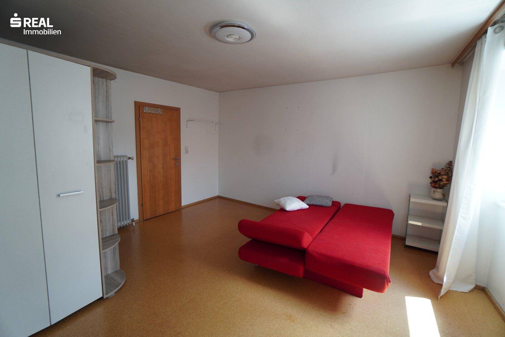 Schlafzimmer mit rotem Schlafsofa, weißem Kleiderschrank und hellem Bodenbelag.