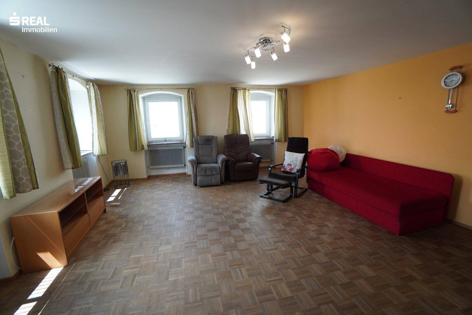 Geräumiges Wohnzimmer mit Parkettboden, orangefarbener Akzentwand und mehreren Fenstern.