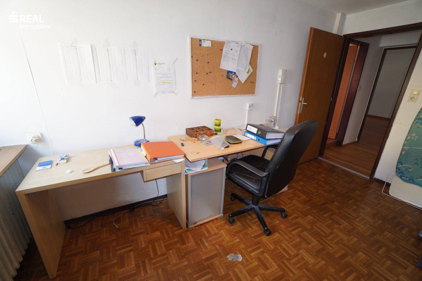 Arbeitszimmer mit großem Holzschreibtisch, Bürostuhl und Korktafel auf Parkettboden.