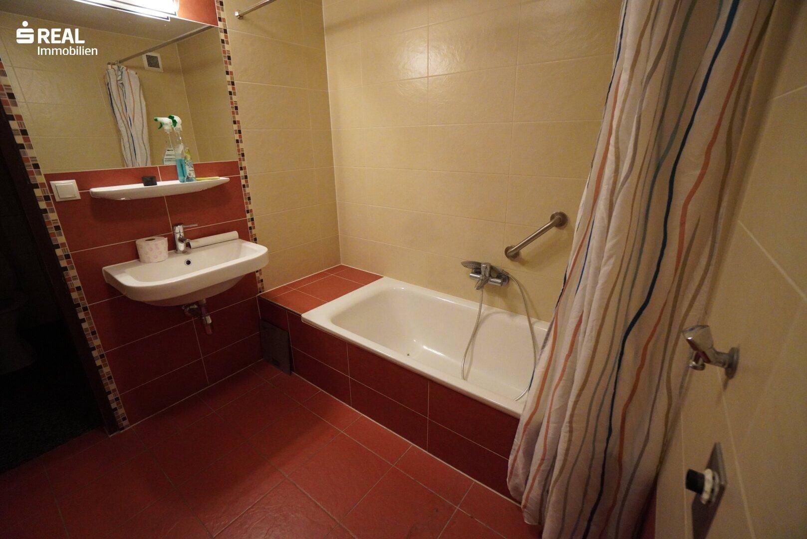 Badezimmer mit weißer Badewanne, Waschbecken und rot-beigen Fliesen, ausgestattet mit Haltegriffen.