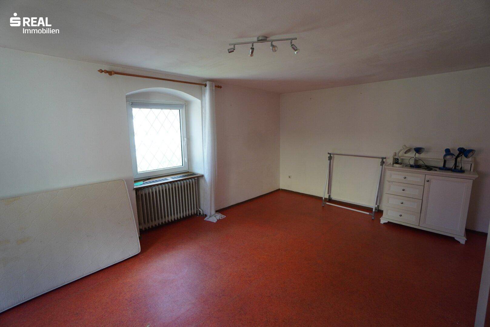 Leerer Raum mit Fenster, Heizkörper und rotbraunem Bodenbelag, bereit zur individuellen Gestaltung.